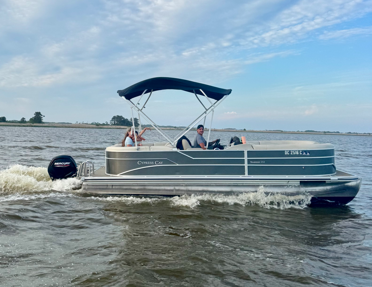 Pontoon boat with canopy on water, two people onboard waving.