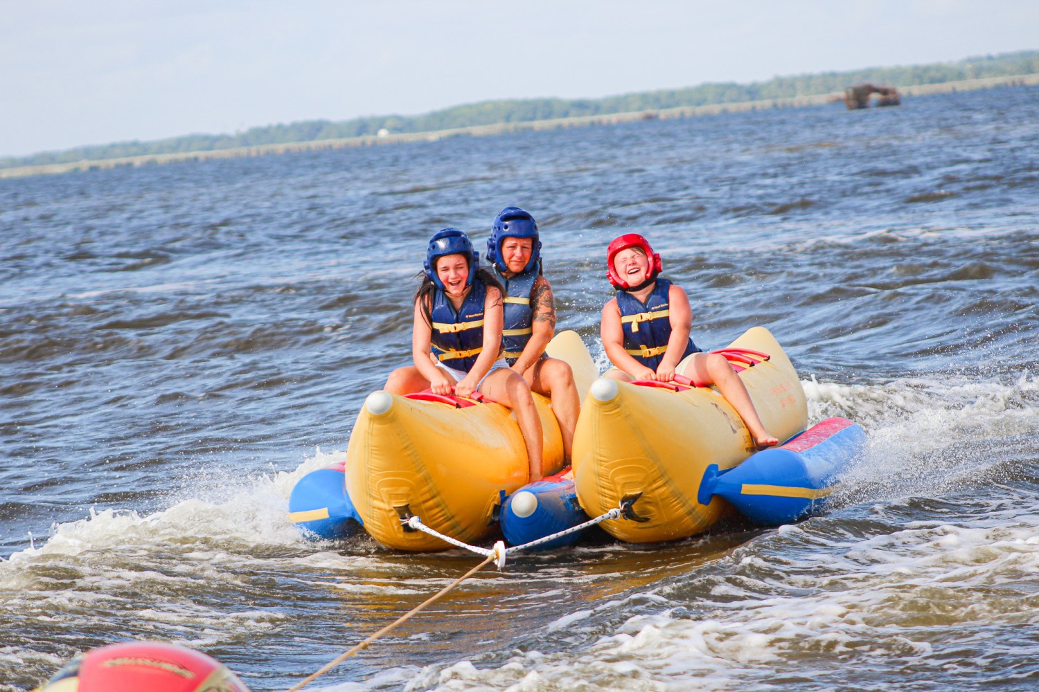 Three people riding a yellow banana boat on water, wearing helmets and life vests.